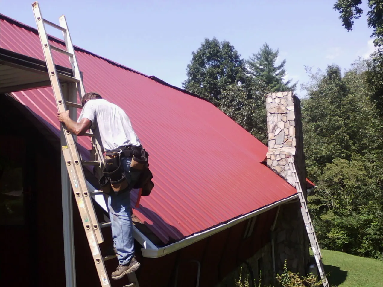 Roofer on a ladder beside a red metal roof and stone chimney — Soffit & Fascia Repair by Roofing Brothers in Anadarko